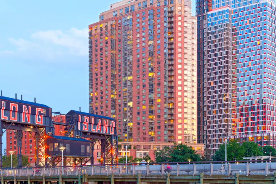 A photo of a bridge reading Long Island City in front of high-rise buildings glittering in a sunset.